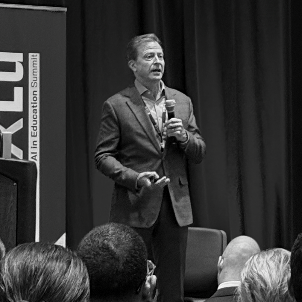 A grayscale image shows a man in a suit speaking at the 'AI in Education Summit.' He holds a microphone, gesturing with his free hand, engaging an unseen audience whose heads are visible in the foreground. The mood is professional and focused.