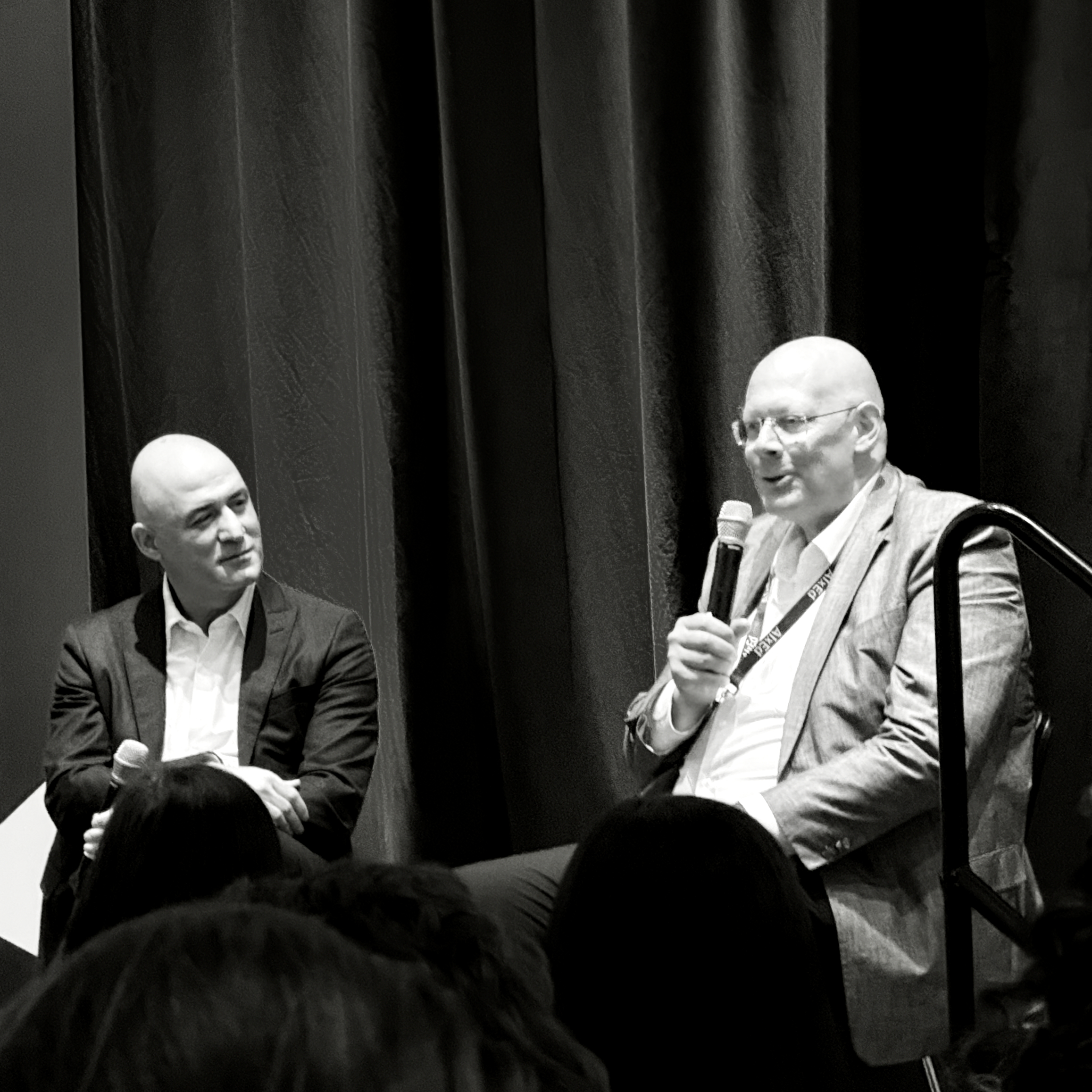 A crisp black-and-white shot shows two bald men on a dimly lit stage. The right man, wearing glasses and a light suit, animatedly speaks into a microphone. The left man, in a dark suit, smiles while attentively listening. Dark drapes backdrop a professional, engaging discussion, with audience heads...