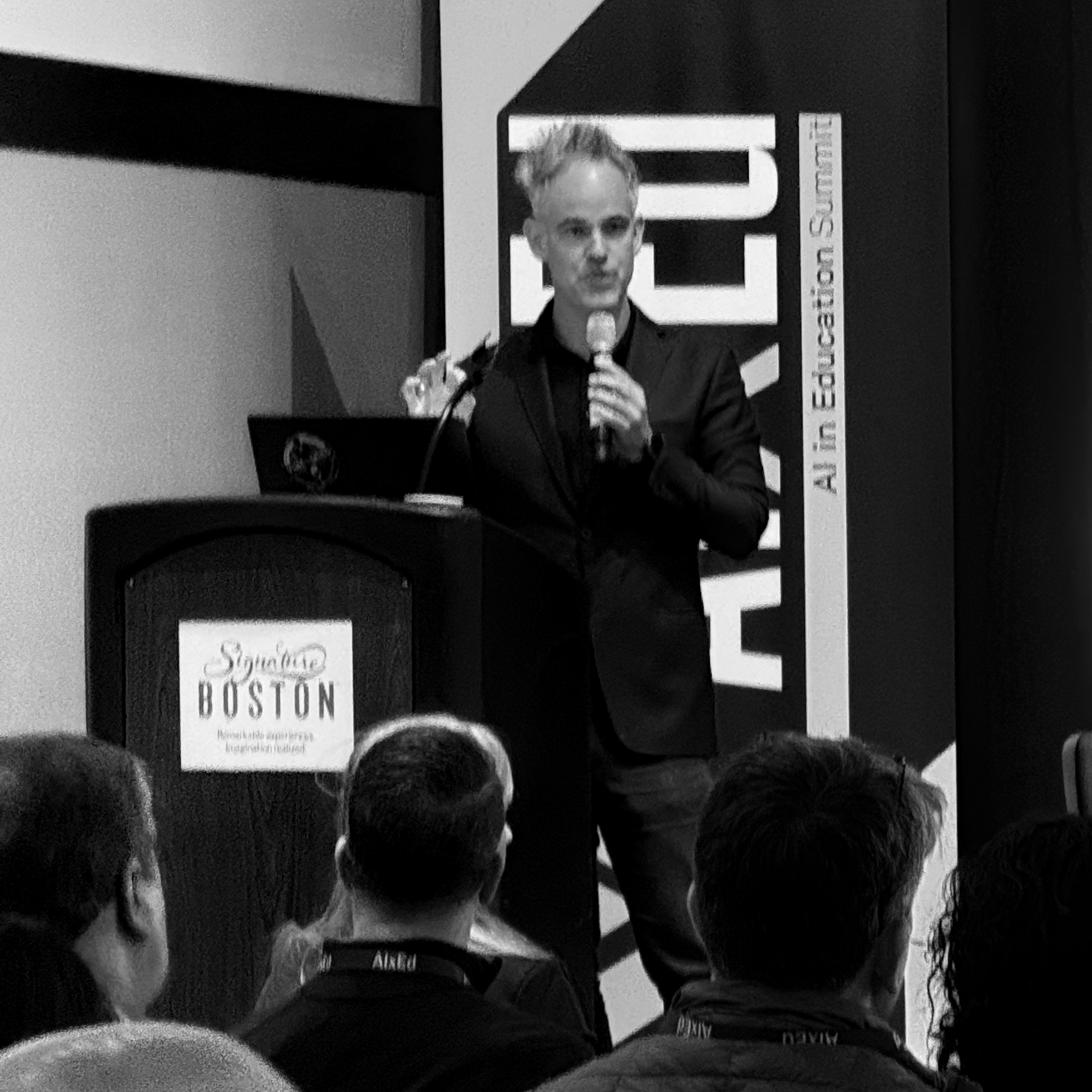 A man with messy hair, in a dark suit, speaks passionately into a microphone from a podium at an 'AI in Education Summit.' The black and white image, shot from behind the audience, creates a focused, professional atmosphere for the engaging presentation.