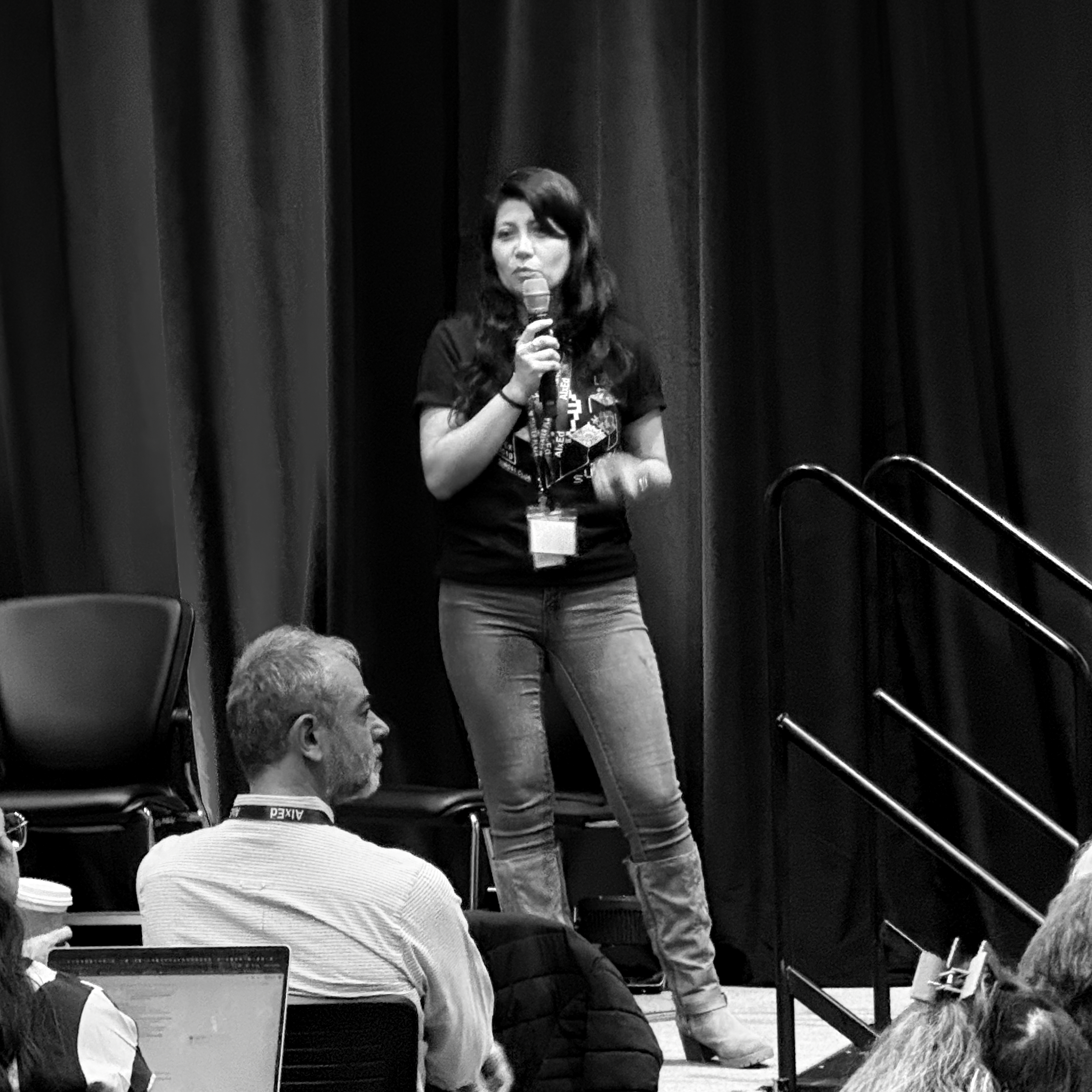 A grayscale image captures a confident woman speaking into a microphone on stage, dressed in a t-shirt, jeans, and boots. Dark curtains backdrop her as audience members, some with laptops, listen intently in the foreground. The scene is a focused presentation.