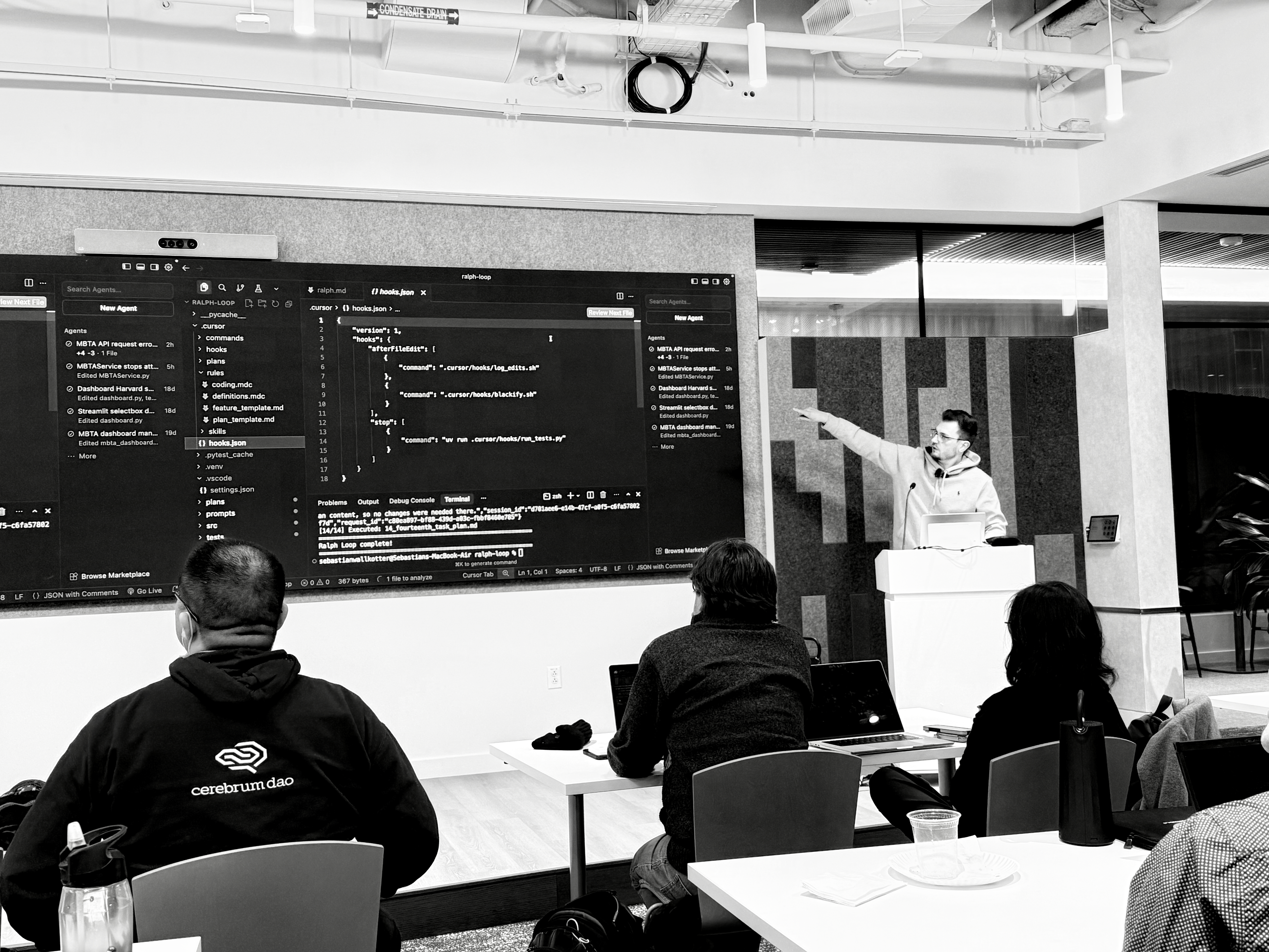 A monochromatic scene captures a speaker pointing emphatically at a large screen displaying 'hooks.json' code, addressing an attentive audience in a modern workspace. The dynamic composition and exposed ceiling elements create an energetic, tech-focused learning mood.