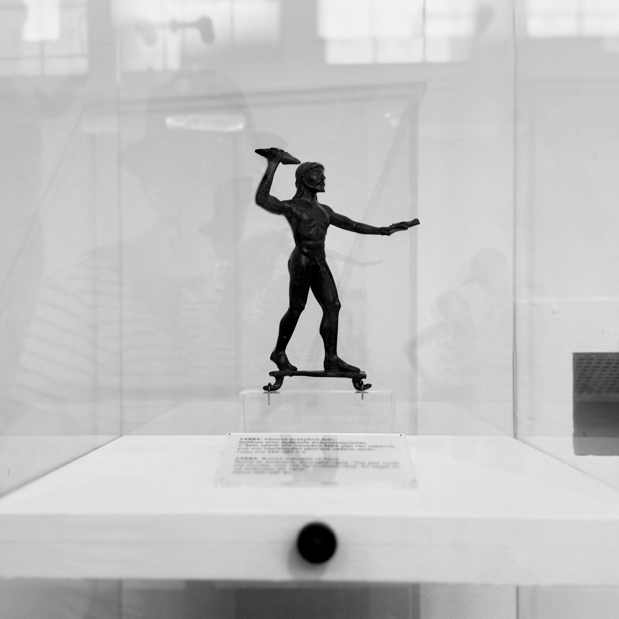 A dark bronze statuette of a powerful, bearded man, poised to hurl a thunderbolt, stands in a grayscale museum display. Set on a white plinth, ethereal reflections of windows and onlookers shimmer behind him, hinting at the present amidst ancient art. His intense pose contrasts with the serene, reflective background.